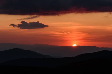 Güneşin dairesel bir diski ile dağların dış hatlarında ve gökyüzündeki aydınlık bulutlarda inanılmaz ateşli bir günbatımı.
