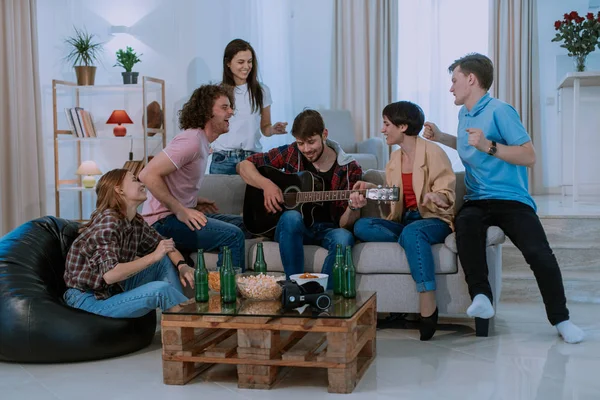 Ett lyckligt och attraktivt företag har en fantastisk tid tillsammans i ett rymligt vardagsrum de sjunger på en gitarr och Dacing klappar händer — Stockfoto