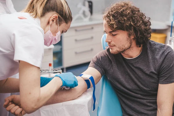 Ärztin und ihre Hilfe machen einen Bluttest bei einem jungen Spender, der das Blut aus der Hand nimmt, einem Mann, der auf dem Behandlungsstuhl in einer modernen Klinik sitzt — Stockfoto