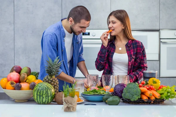 Una joven pareja está preparando verduras y frutas para el batido, y la dama está disfrutando de la zanahoria —  Fotos de Stock