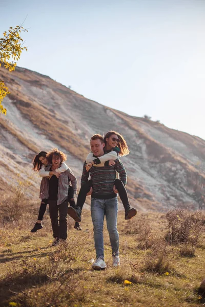 Dos jóvenes pareja pasar un buen rato juntos en la naturaleza, chicos sosteniendo en su espalda a sus novias y caminando por el campo sonriendo y con un buen humor —  Fotos de Stock