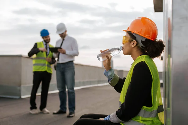 En el sitio de construcción hermosa sonriente ingeniero africano bebiendo un poco de agua de la botella en la azotea del edificio que llevaba casco de seguridad y gafas de seguridad amarillas —  Fotos de Stock