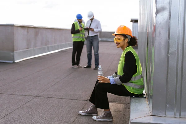 Carismatico ingegnere donna africana sorridente carino bere un po 'd'acqua sul tetto del cantiere sfondo i suoi colleghi analizzando il piano di costruzione — Foto Stock