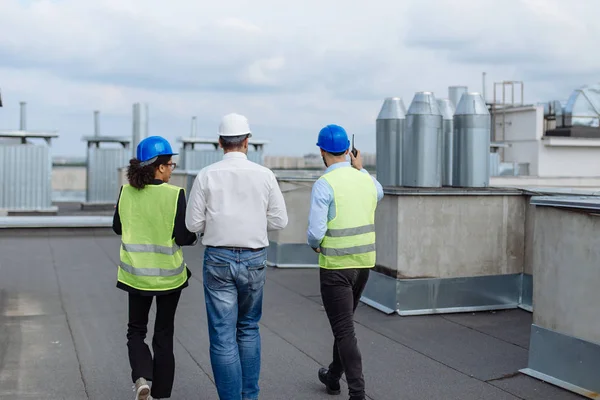 Tirando vídeo do telhado traseiro do canteiro de obras um arquiteto e dois engenheiros multiétnicos analisando o plano de construção enquanto caminham eles vestindo capacetes de segurança . — Fotografia de Stock