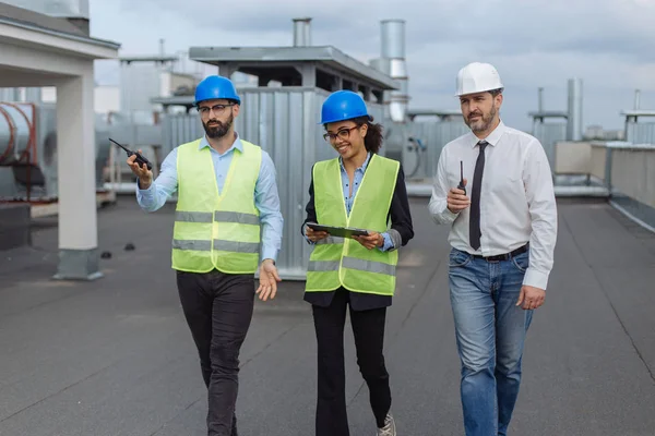 Dois arquitetos homens e sua ajuda mulher africana analisando o plano de canteiro de obras andando no telhado da construção segurando um tablet eles têm uma discussão concentrada . — Fotografia de Stock