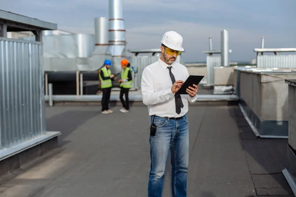 Sul tetto del cantiere uomo ingegnere fare una conversazione con alcuni lavoratori utilizzando il tablet due lavoratori sono visti sullo sfondo — Foto Stock