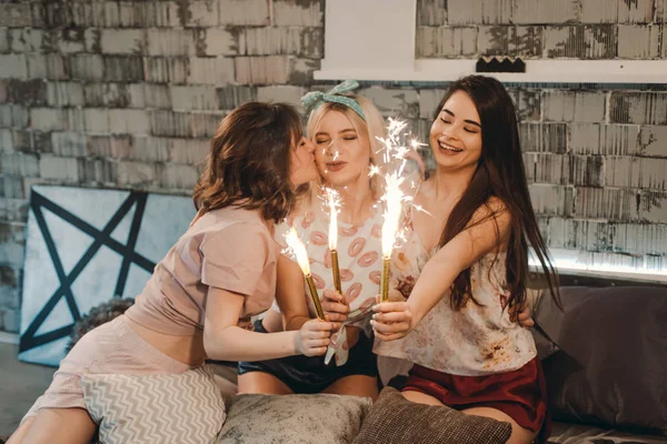 Las damas multiétnicas en la noche de pijamas hacen que la noche sea más divertida en un dormitorio moderno usando fuegos artificiales brillantes, se besaron en la mejilla —  Fotos de Stock