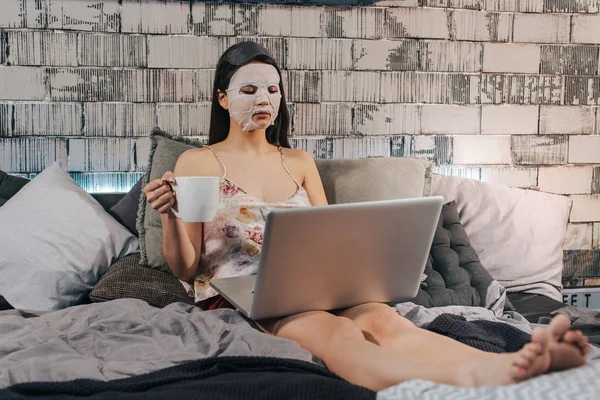 Rutina de belleza matutina para una mujer de negocios acostada en la cama con una máscara facial en su computadora portátil mientras bebe café . —  Fotos de Stock