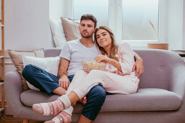 Watching tv young couple in the morning sitting on the sofa and eating a big plate of popcorn wearing a pajamas