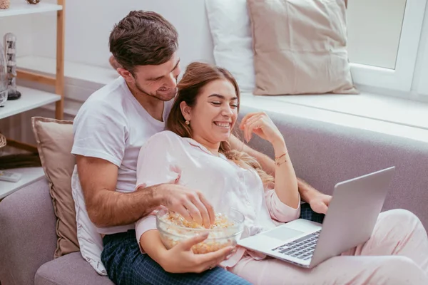 Watching tv young couple in the morning sitting on the sofa and eating a big plate of popcorn wearing a pajamas