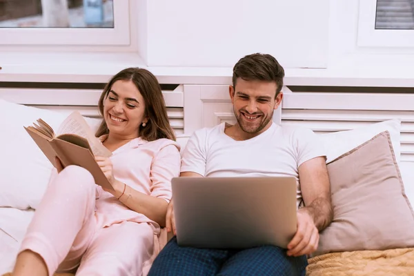 Morning routine for a couple in bed , woman reading a book and her partner are looking on the laptop