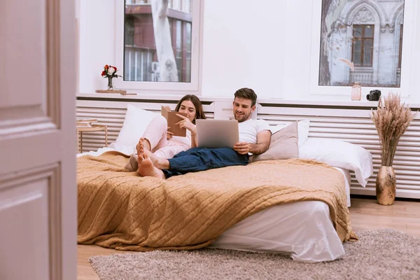 El hombre y su novia por la mañana en la cama, diseño acogedor, libro de lectura y mirando en el ordenador portátil —  Fotos de Stock