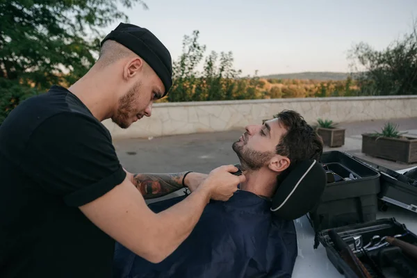 Barber shaves beard of client man on chair an open air