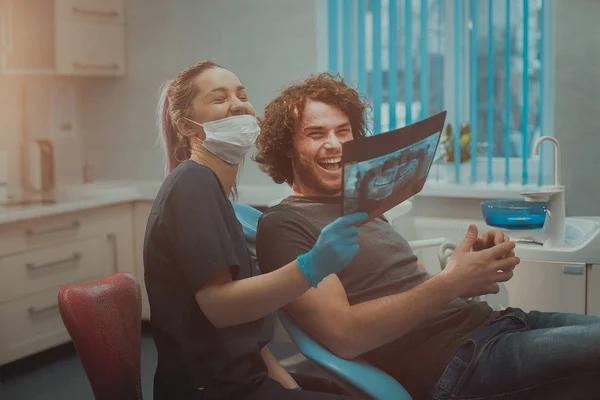 Young dentist with blue gloves and mask is amused with his patient, the result of the x-ray of teeth — Stock Photo, Image