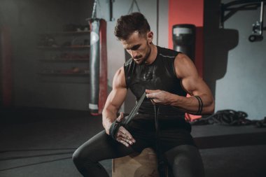 Portre konsantre acımasız atletik adam jimnastik sınıfında sandalyede otururken profesyonel bandajlar kullanarak ellerini sarar