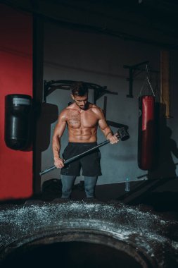 Sert egzersizler güçlü atletik adam, güçlü vücut gücüyle dolu büyük bir lastiğe vurmak için hummer kullanıyor.