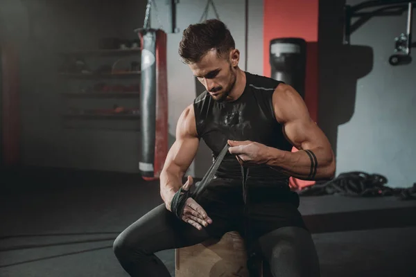 Retrato concentrado tipo atlético brutal envuelve las manos usando las vendas profesionales mientras está sentado en la silla en la clase de gimnasia — Foto de Stock