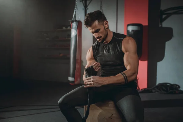 Retrato concentrado tipo atlético brutal envuelve las manos usando las vendas profesionales mientras está sentado en la silla en la clase de gimnasia — Foto de Stock