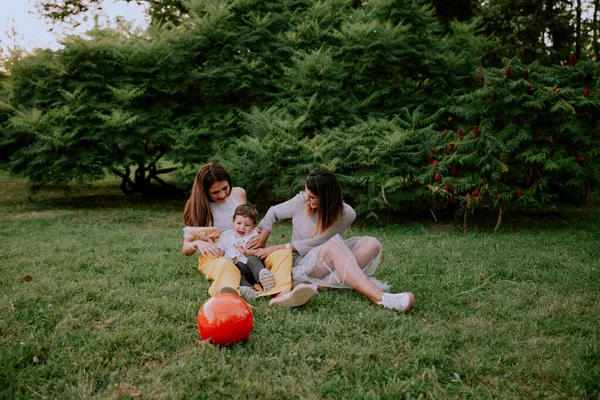 Muy lindo niño y dos sonrientes señoras atractivas grandes disfrutando del tiempo en el parque acostado en la hierba —  Fotos de Stock