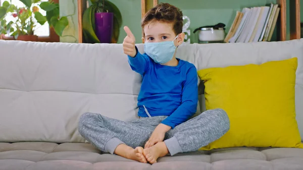 Yeni Covid-19 küçük çocuk oturma odasındaki kanepede oturuyor koruyucu maske takıyor kameraya bakıyor ve evde güvende ve sağlıklı olmak için başparmağını kaldırıyor.