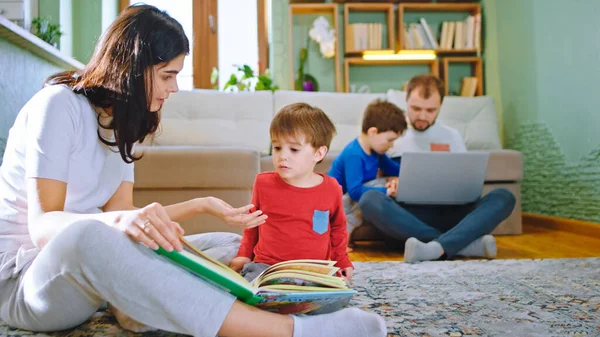 Duży młody rodzina spędzają dzień w domu mama czyta historię z jej syn tata z innym dzieckiem robi niektóre działania z laptopa — Zdjęcie stockowe