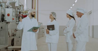 Yiyecek endüstrisi mühendisi elinde bir tablet tutuyor ve işçilerine makineyle ilgili talimatları anlatıyor. Korunmak için özel kıyafetler giyiyorlar.