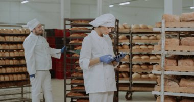 Güzel bayan fırıncı tabletine bazı notlar ve fırın endüstrisindeki raflardan fırınlanmış ekmek fotoğrafları al.