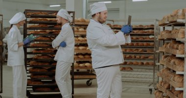 Ana fırıncı yeni bir teknoloji tableti kullanarak arka planda konuşan diğer iki işçinin raflardan taze pişmiş ekmek fotoğraflarını çekiyor.