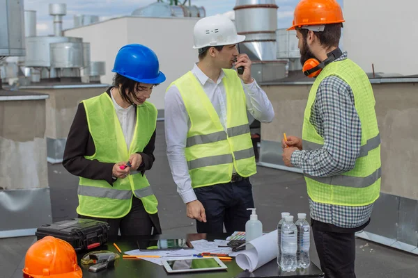 Gruppo multietnico di lavoratori architetto e due ingegneri hanno una conversazione sul tetto dell'edificio analizzando cantiere e parlando attraverso la razione con altri lavoratori — Foto Stock