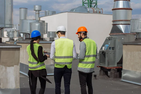 Scattando video dal tetto posteriore del cantiere un architetto e due ingegneri multietnici analizzano il piano di costruzione mentre camminano indossano caschi di sicurezza — Foto Stock