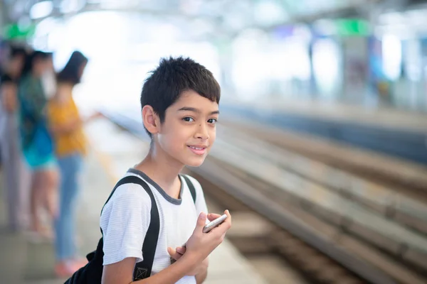 手にモバイルでプラットフォーム上で列車を待っている小さな学生の男の子 — ストック写真