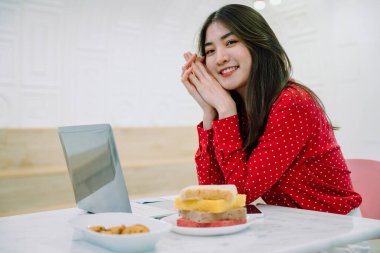 Güzel Asyalı, uzun siyah saçlı, kırmızı tişörtlü, dizüstü bilgisayarlı ve masada atıştırmalıklarla çalışan bir kadın..