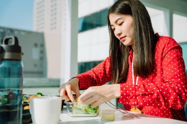 Kırmızı tişörtlü güzel Asyalı kadın ofis yerinde cam kasede salata yiyor..