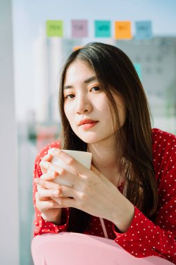 Sabahları elinde kahve fincanı tutan kırmızı tişörtlü güzel Asyalı genç kadının portresini yakından çek. Şehir görünümü arka planı.