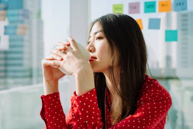 Sabahları elinde kahve fincanı tutan kırmızı tişörtlü güzel Asyalı genç kadının portresini yakından çek. Şehir görünümü arka planı.