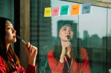 Beautiful young asian thai businesswoman in red shirt reading pose it or sticky paper note on glass wall. clipart