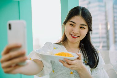 Neşeli güzel Asyalı Taylandlı kadın akıllı telefon kullanarak mango yapışkan pirinçle selfie çekiyor..