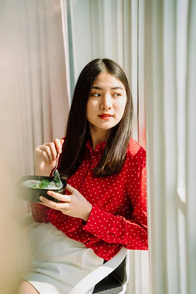 Beautiful asian thai long dark hair woman in red shirt enjoy eating healthy food, sitting on a chair on office room. Working at home. Work from home.