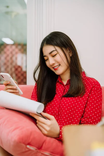 Beautiful asian thai long dark hair woman in red shirt sitting on pink sofa couch using smartphone to make contact with customer and checking list on paper work. Work from home. Working at home.