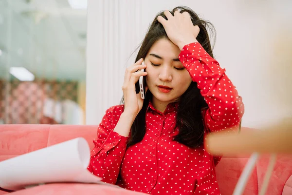 Beautiful asian thai long dark hair woman in red shirt sitting on pink sofa couch using smartphone to make contact with customer and checking list on paper work. Work from home. Working at home.