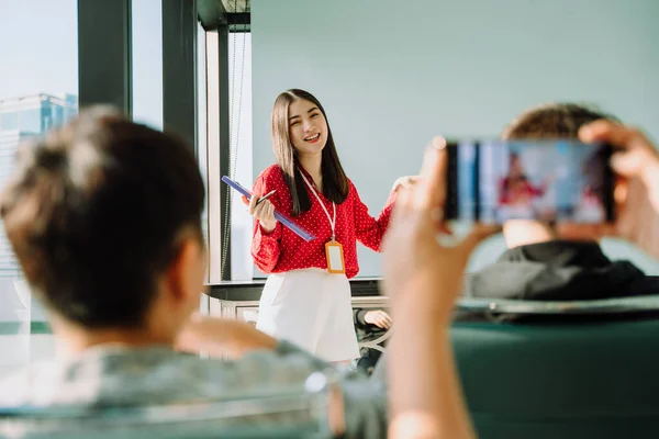 Beautiful young asian thai businesswoman in red shirt giving present in front of the room. People taking picture or record the video of the presentation with smartphone.