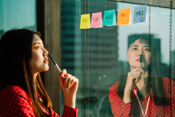 Beautiful young asian thai businesswoman in red shirt reading pose it or sticky paper note on glass wall.