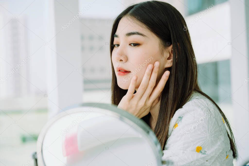 Beautiful asian young thai woman putting cream make up on face in front of the mirror.