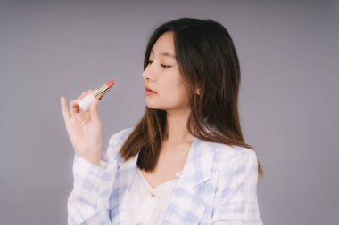 Gri arka planda izole edilmiş ruj kozmetiği olan beyaz moda gömlekli güzel Asyalı Taylandlı kadın. Kozmetik hastası bir kadın. Uzun siyah saçlı..