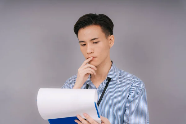 Handsome thai businessman in blue collar shirt checking business report on clipboard with thoughtful expression. Isolated over grey background.