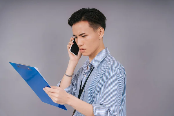 Handsome thai businessman in blue collar shirt checking report on clipboard and talking on smartphone seriously. Isolated over grey background.