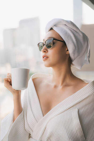 Портрет крупным планом - молодая женщина в белом халате и солнцезащитных очках, пьющая из чашки кофе на фоне города
.