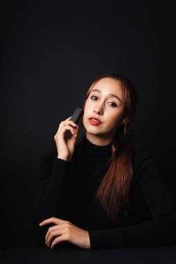 Siyah kazak giyen güzel bir genç kadının yakın plan portresi koyu arkaplan üzerine izole edilmiş. Karanlıktaki güzellik. Elinde siyah ruj kutusu var..