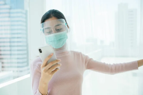 新しい普通の生活 自宅でスマートフォンを使用して顔の盾とマスクを身に着けているアジアのタイ人女性 — ストック写真
