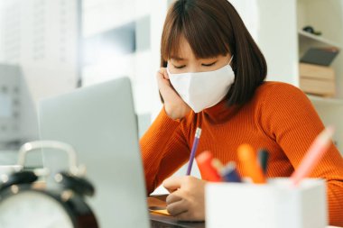Genç, güzel Asyalı Taylandlı kısa saçlı, beyaz cerrahi maske takan, laptopla evde ders çalışan bir kadın..